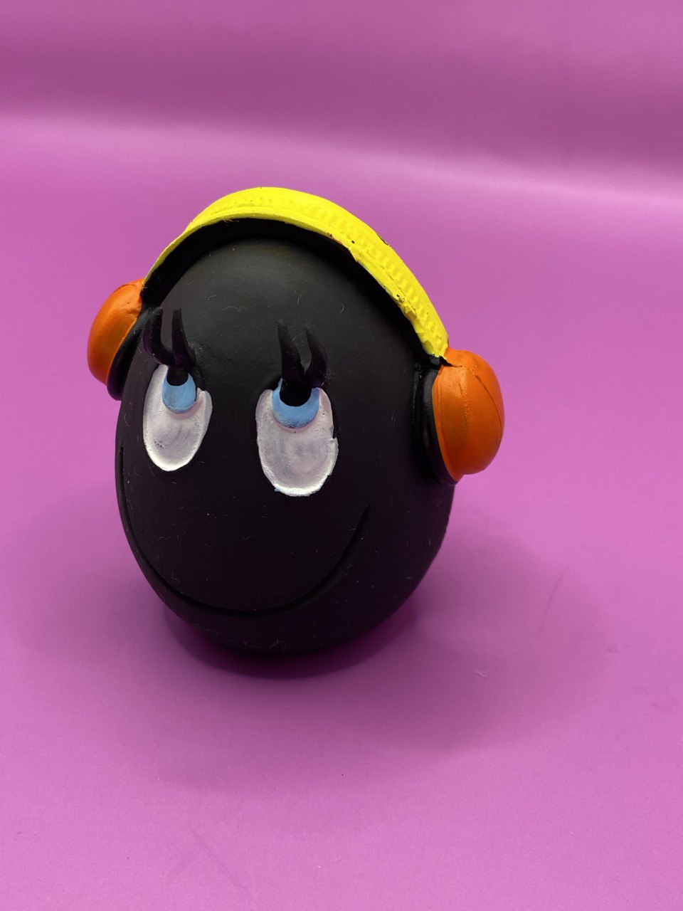 Huevo DJ de Látex de LANCO para perros de color negro con auriculares donde la parte de la diadema es amarilla y los altavoces son naranjas. Sus ojos son azules con la pupila en negro y están mirando hacia arriba como con cara de bueno, también  tiene una línea en la boca sonriendo 3